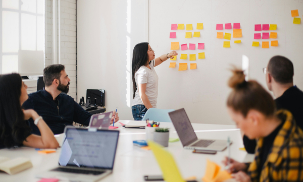 People sitting around a table, with laptops, watching as a woman organises colourful post-it notes